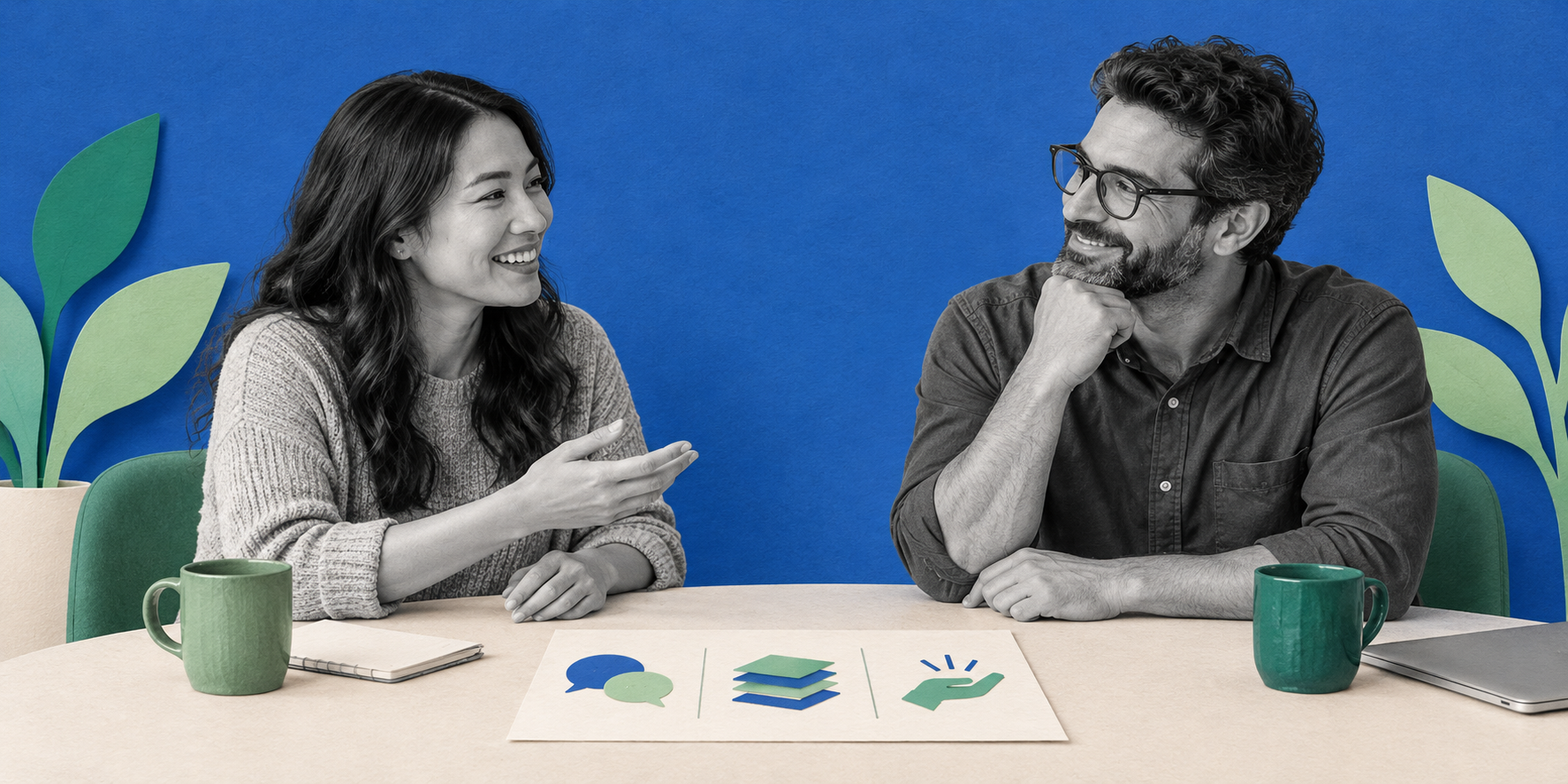 Two coworkers smiling and talking across a table with green mugs